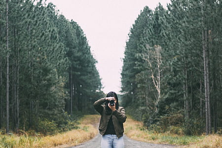 woman holding camera standing on road between trees at daytime thumbnail