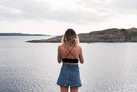 woman standing in front of body of water and mountain under cloudy sky thumbnail