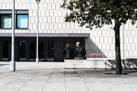 two men sitting on concrete bench in front of white painted building thumbnail