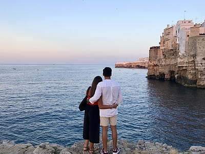 Man in White Dress Shirt Standing Beside the Woman in Black and Red Dress While Watching the Blue Calm Water Near Brown Concrete Buildings Under White and Blue Sky at Daytime thumbnail