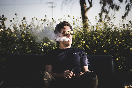sitting man wearing Aviator-style sunglasses blowing smoke while holding box mod and sitting on bench behind flowering plants thumbnail