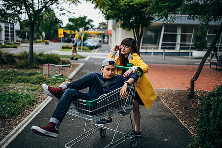 man riding grey shopping cart in front of woman thumbnail