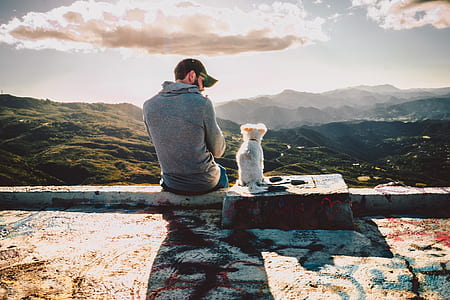 man in gray shirt sitting beside white puppy under cloudy sky thumbnail