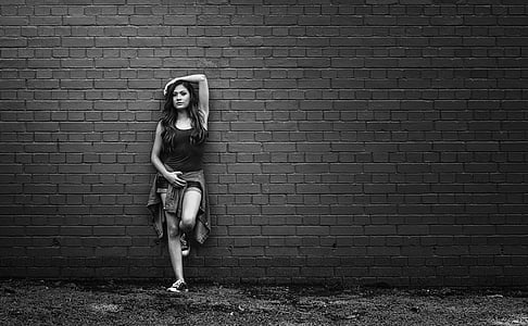 grayscale photo of woman wearing tank top and shorts with jacket on waist leaning on wall thumbnail