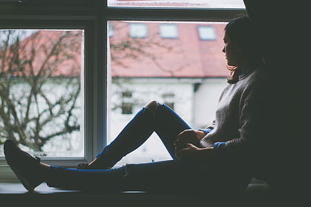 woman in distressed denim jeans and sweater leaning beside wall post while watching outside the window thumbnail