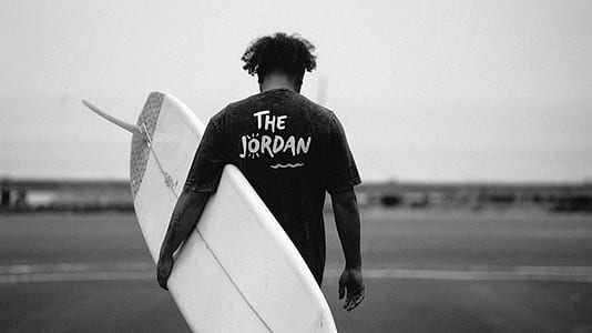 grayscale photo of man carrying surfboard thumbnail