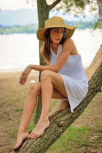 woman sitting on tree branch wearing straw hat near body of water at daytime thumbnail