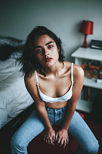 woman in white bra and blue jeans sitting beside bed thumbnail