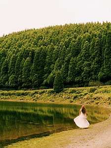 woman standing near body of water and trees while wearing white dress during daytime thumbnail