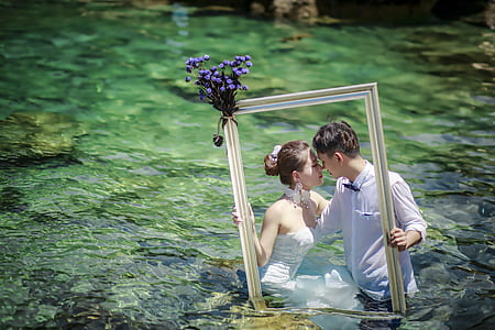 couple in wedding dress holding photo frame standing on body of water thumbnail