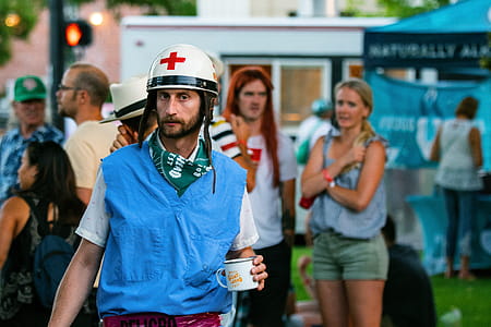 Man Holding Cup in a White Helmet with a Red Cross Near Group of People thumbnail