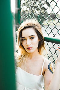 woman wears white sleeveless top leaning on green fence thumbnail