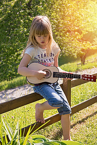 girl wearing white t-shirt and blue denim shorts playing brown ukulele leaning on brown wooden fence during daytime thumbnail