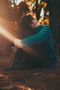 Selective Focus Photography of Woman in Green Jacket and Black Pants Sitting on Field Surrounded With Trees thumbnail