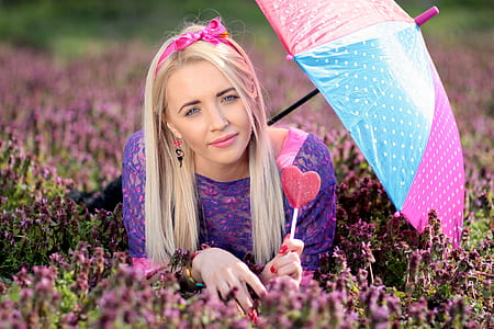 woman prone lying on flower field under shade of umbrella while holding heart-shaped pink lollipop thumbnail
