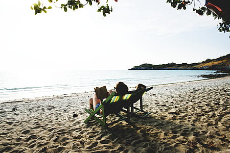 person reading book while lying on lounge chair near ocean during daytime thumbnail