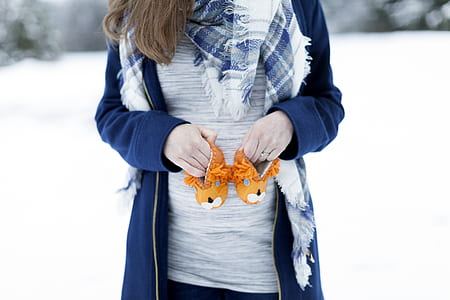 woman holding pair of baby's orange winter boots while standing on snow covered land thumbnail