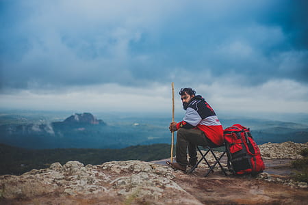 man sitting on folding chair beside red hiking backpack near view of mountain at daytime thumbnail