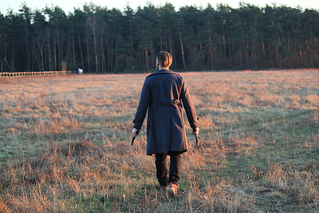 man holding two guns and walking on grass field during daytime thumbnail