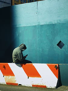 person wearing green hoodie sitting on white and orange concrete road barricade during daytime thumbnail