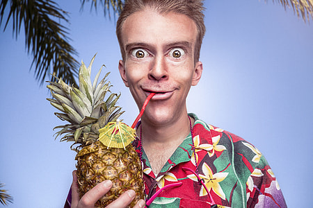 man in red, yellow, and green flora button-up shirt holding pineapple under blue sky thumbnail