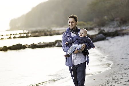 man carrying baby near seashore during daytime thumbnail