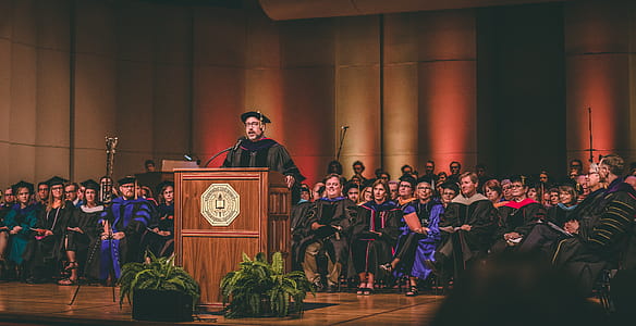 Man Standing at a Center of Stages on Podium Surrounded by People thumbnail