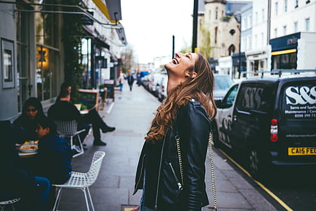 woman in black leather jacket laughing on road side near cars parked on gray asphalt road during daytime thumbnail
