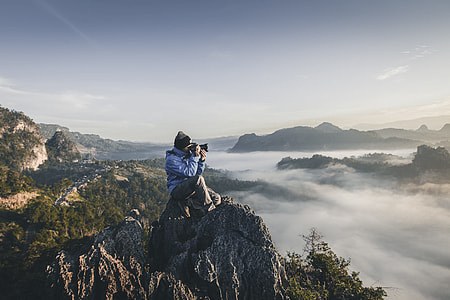 man sitting on mountain peak holding camera thumbnail