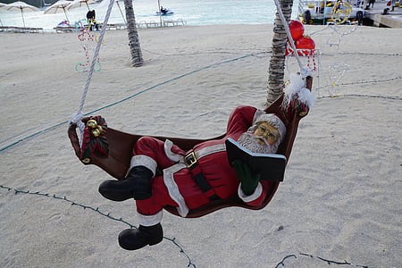 Santa Claus lying on brown hammock reading book near body of water at daytime thumbnail
