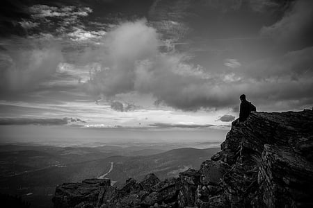 man sitting on rocky cliff looking at mountains in grayscale photography thumbnail
