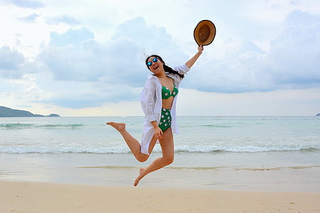 woman wearing bikini holding hat while jumping in seashore thumbnail