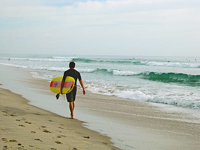 man in black shirt and shorts carrying a yellow surfboard on beach thumbnail