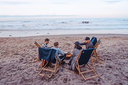 four people sitting on adirondack chairs facing in fire pit on seashore thumbnail