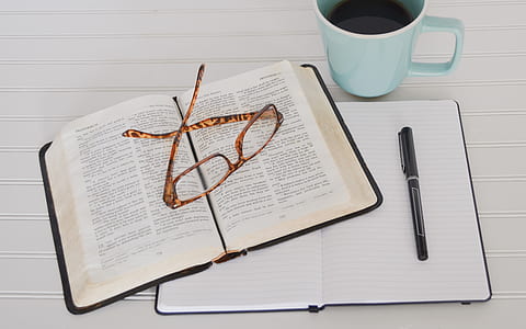 flat lay photography of open bible, notebook, pen, eyeglasses and mug of coffee thumbnail