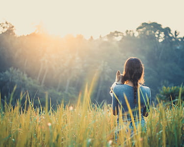 woman carrying her pet in the middle of grass facing trees at daytime thumbnail