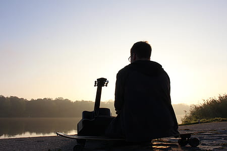 silhouette photo of man sitting beside guitar facing at the lake across forest at daytime thumbnail