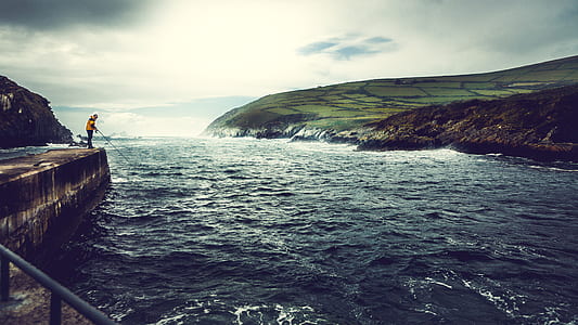 person standing on rock while holding fishing rod near body of water at daytime thumbnail
