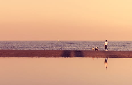 Man Standing Near Seashore With Boat in Distance Under Cloudy Sky during Daytime thumbnail