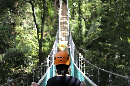 woman wearing orange helmet standing on brown wooden hanging bridge during daytime thumbnail