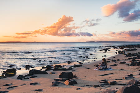 woman sitting on blanket at shore while watching the sea under white cloudy sky during golden hour thumbnail