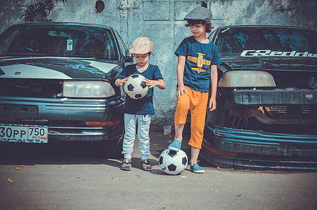Two Boys Playing Soccer Ball Beside Cars thumbnail