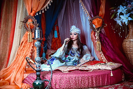woman wearing blue long-sleeved dress and pagri hat sitting on bed smoking hookah thumbnail