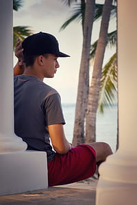 man in gray t-shirt sitting near concrete post in a distance of coconut trees thumbnail