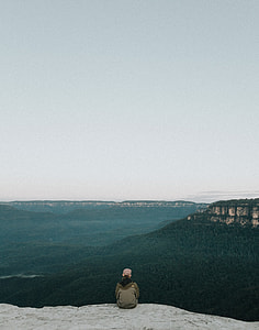 person wearing grey jacket sitting on edge of cliff facing mountains thumbnail