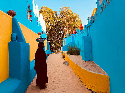 silhouette of person with basket on head standing in between blue and yellow buildings during daytime thumbnail