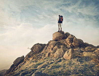 man carrying hiking bag standing on rocky cliff during daytime thumbnail