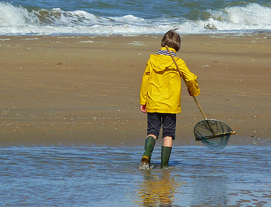boy wearing yellow rain coat and blue capri pants with pair of green rain boots on body of water holding net thumbnail
