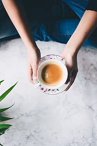 person holding white ceramic cup filled with brown liquid on white and pink floral saucer thumbnail