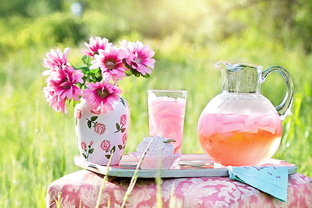 clear glass of pitcher with juice near glass full of juice and pink flowers in vase on table thumbnail